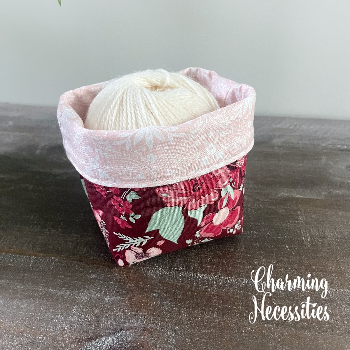 Floral yarn bowl with a ball of yarn on a wooden surface, branded 'Charming Necessities'.