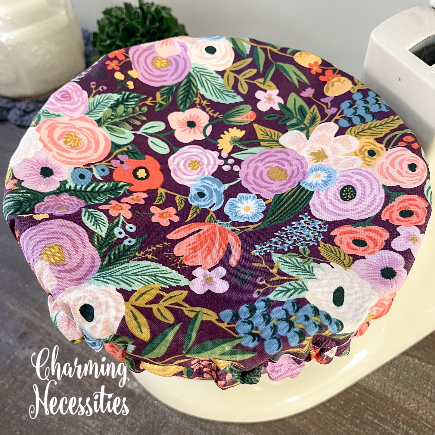 Floral-patterned bowl cover on a white surface with 'Charming Necessities' branding