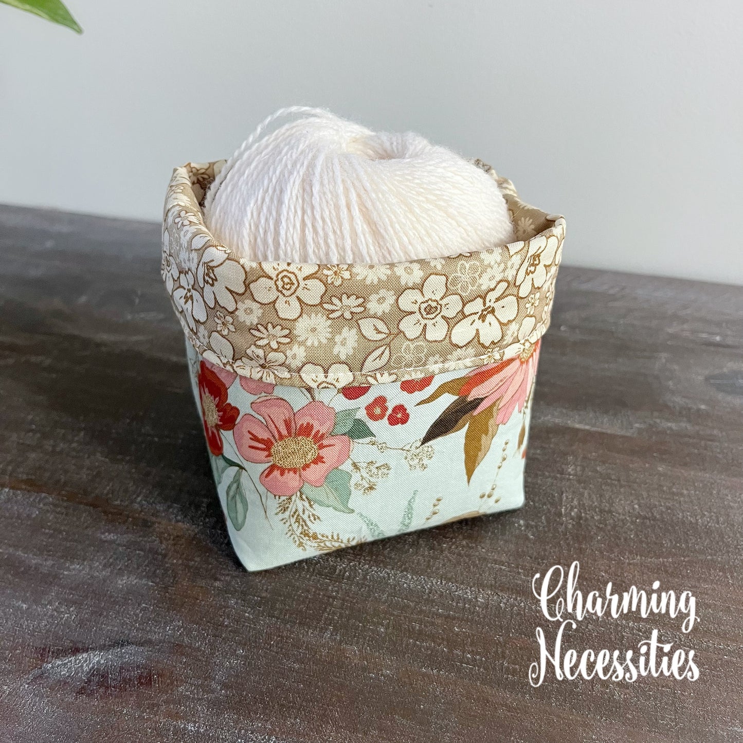Floral-patterned yarn bowl with yarn inside on a wooden surface, featuring 'Charming Necessities' branding.