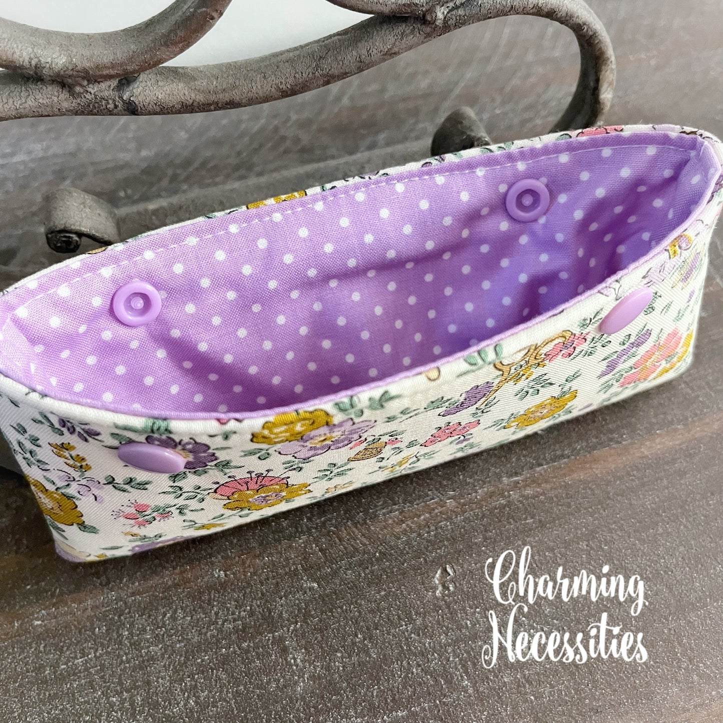 Needle holder pouch with floral pattern and purple interior on a wooden surface, branded 'Charming Necessities'