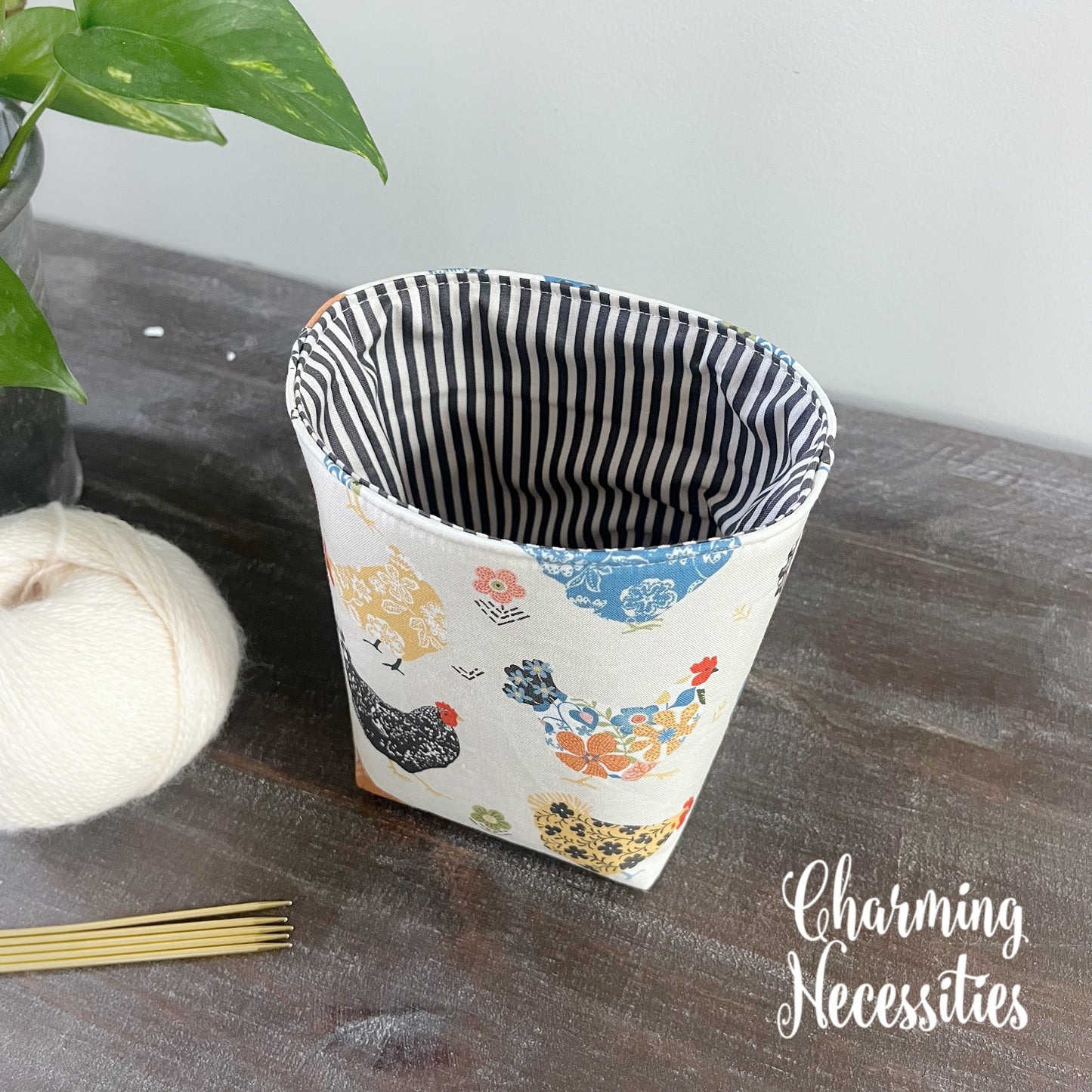 yarn bowl with chicken pattern on a wooden surface, next to yarn and knitting needles, with 'Charming Necessities' branding.