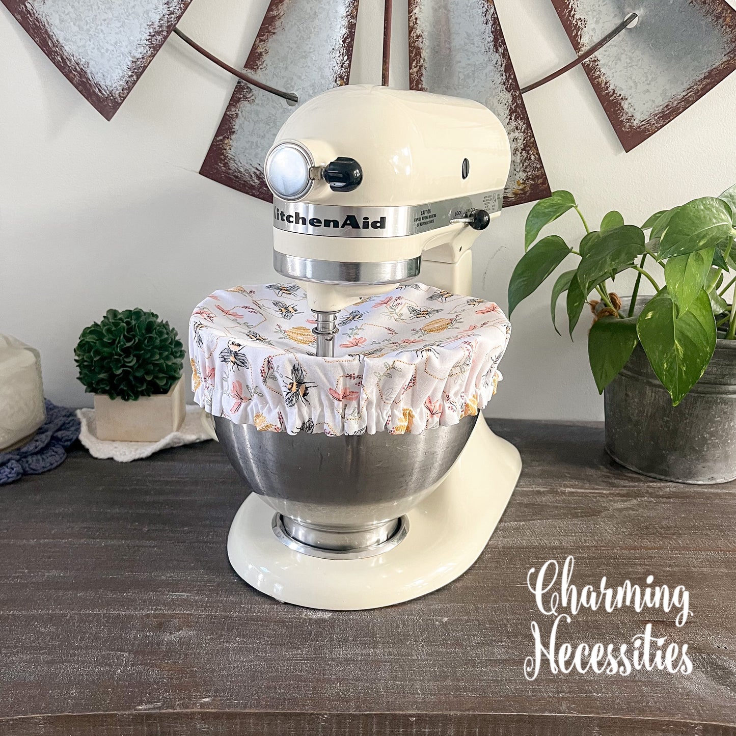 KitchenAid mixer with a bee themed cover on a wooden surface, surrounded by decorative items.
