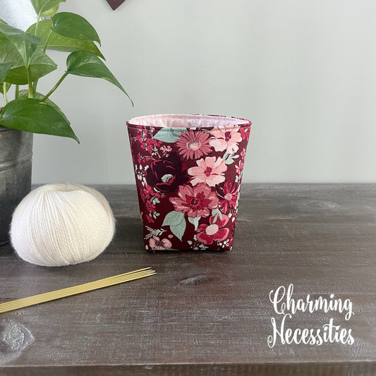 dark red Floral-patterned fabric yarn basket on a wooden surface with yarn and knitting needles, branded 'Charming Necessities'.