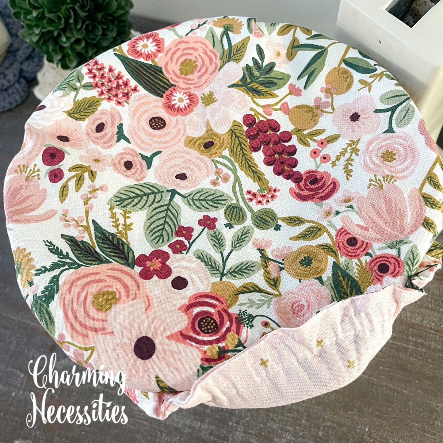 Floral-patterned bowl cover showing contrasting fabric on a wooden surface, featuring the brand 'Charming Necessities'.