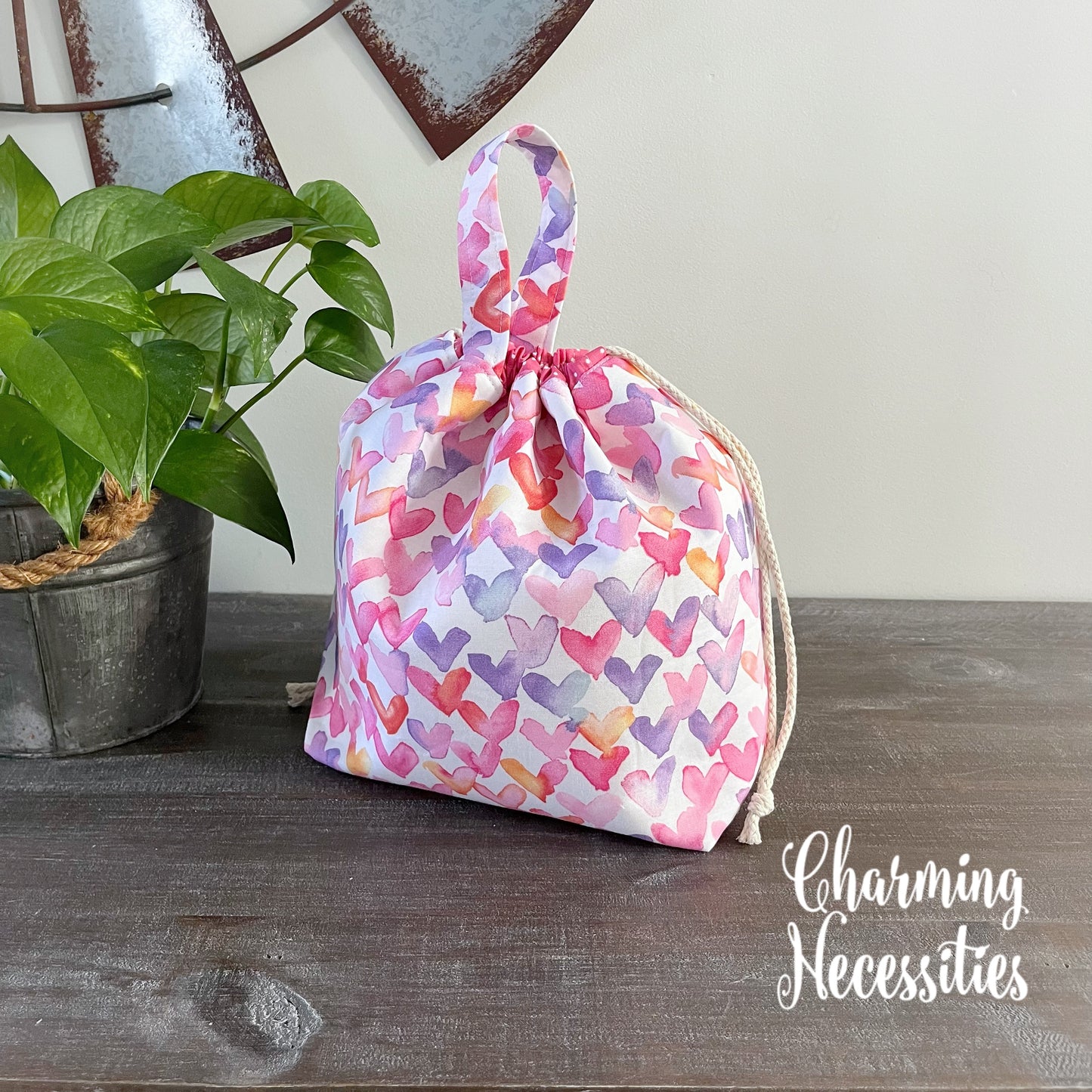 Drawstring bag with heart pattern on a wooden surface next to a plant, with 'Charming Necessities' branding.