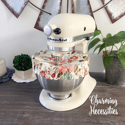 KitchenAid mixer with a floral cover on a wooden surface, surrounded by decorative items.