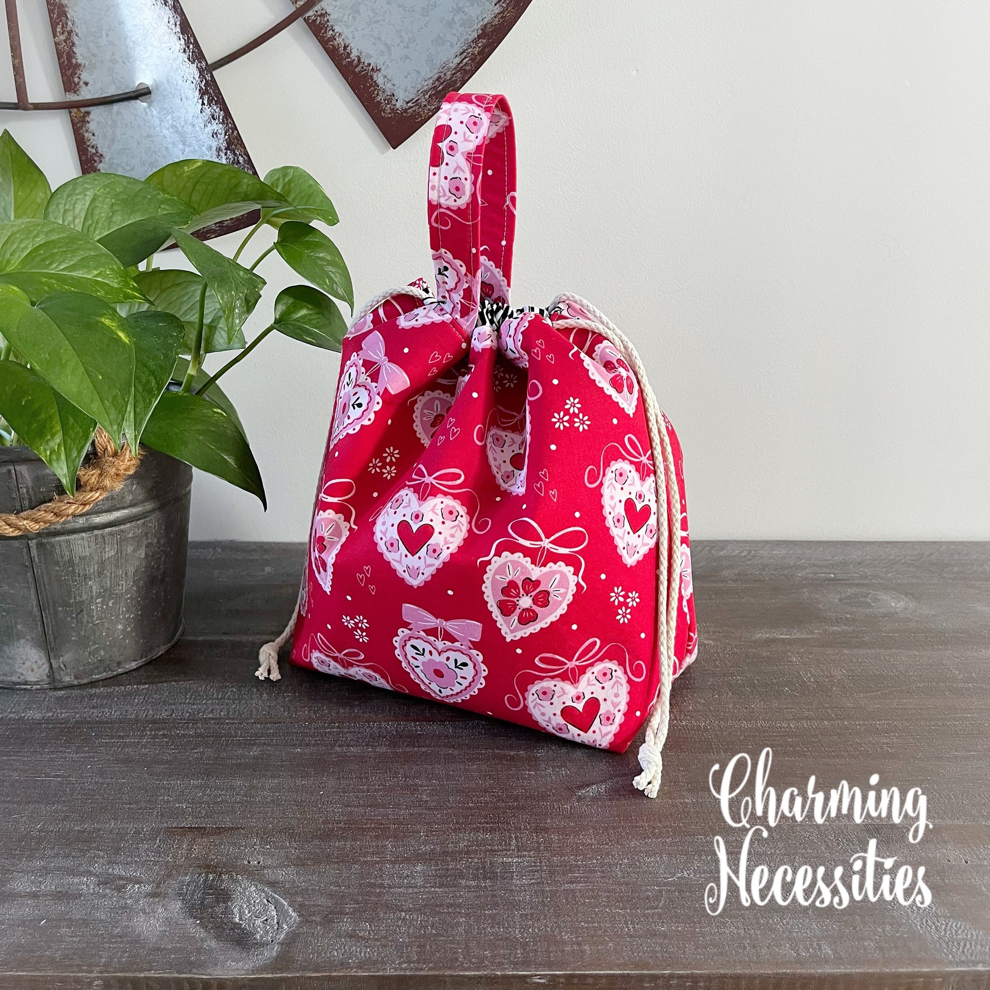 Red drawstring bag with heart patterns on a wooden surface, next to a plant.