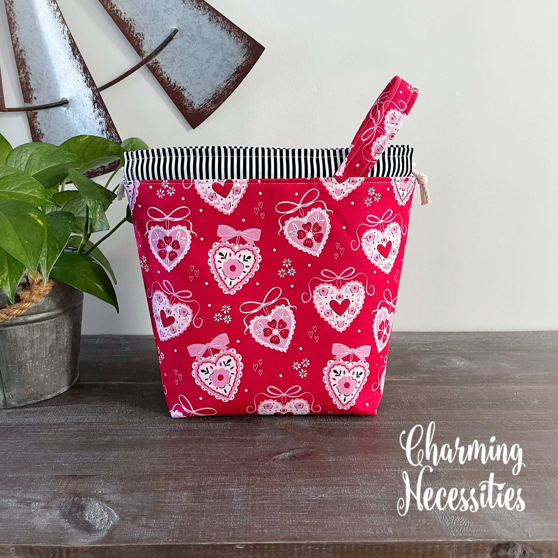 Red fabric drawstring bag with heart pattern on a wooden surface, featuring the brand 'Charming Necessities'.