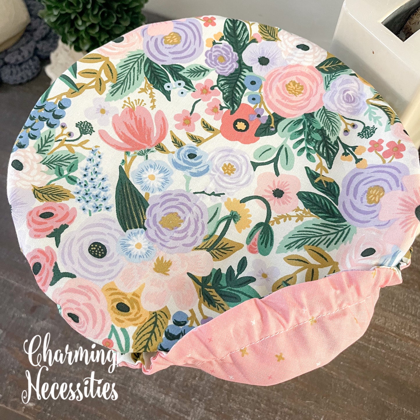 Floral-patterned bowl cover with a pink border on a wooden surface, branded 'Charming Necessities'.