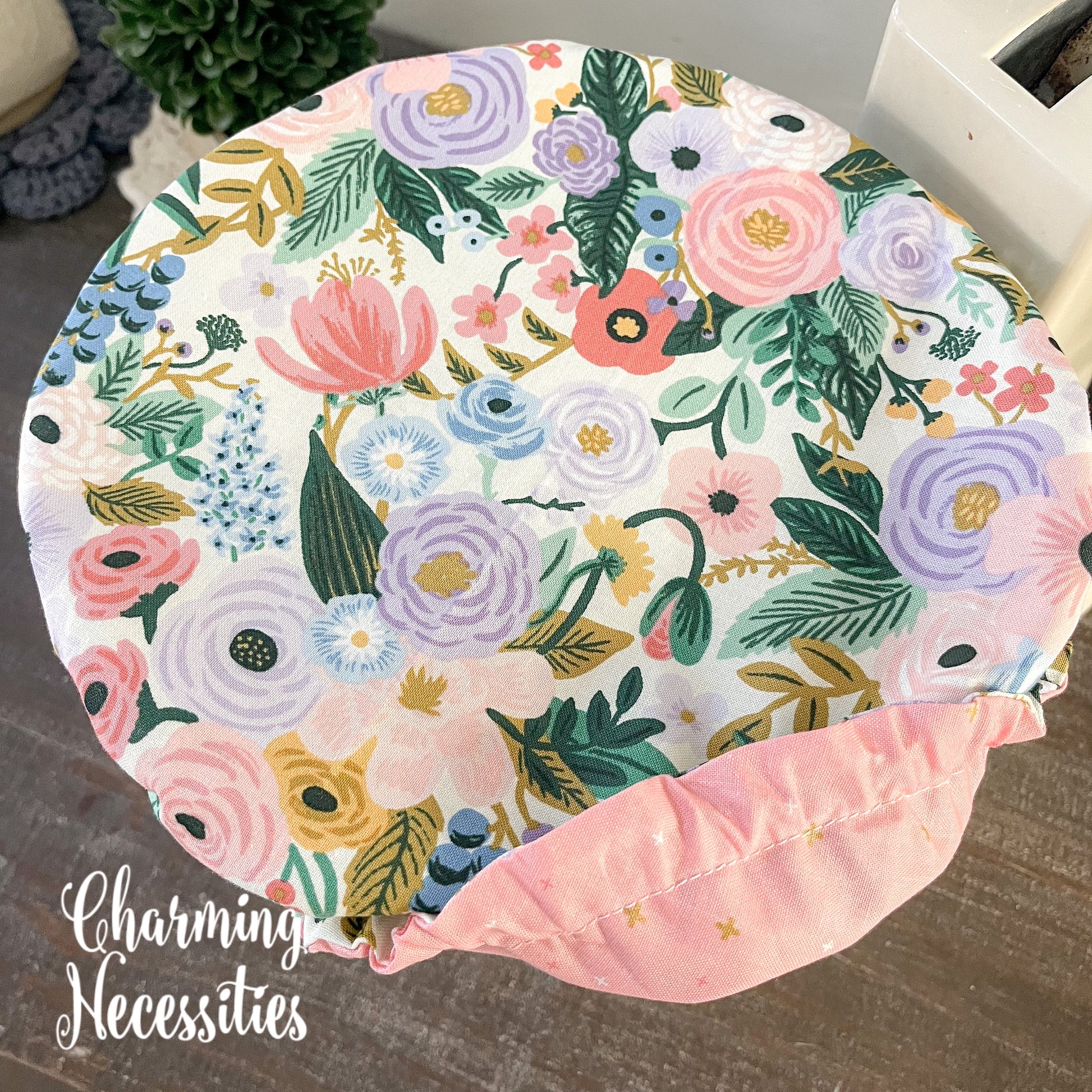 Floral-patterned bowl cover with a pink border on a wooden surface, branded 'Charming Necessities'.