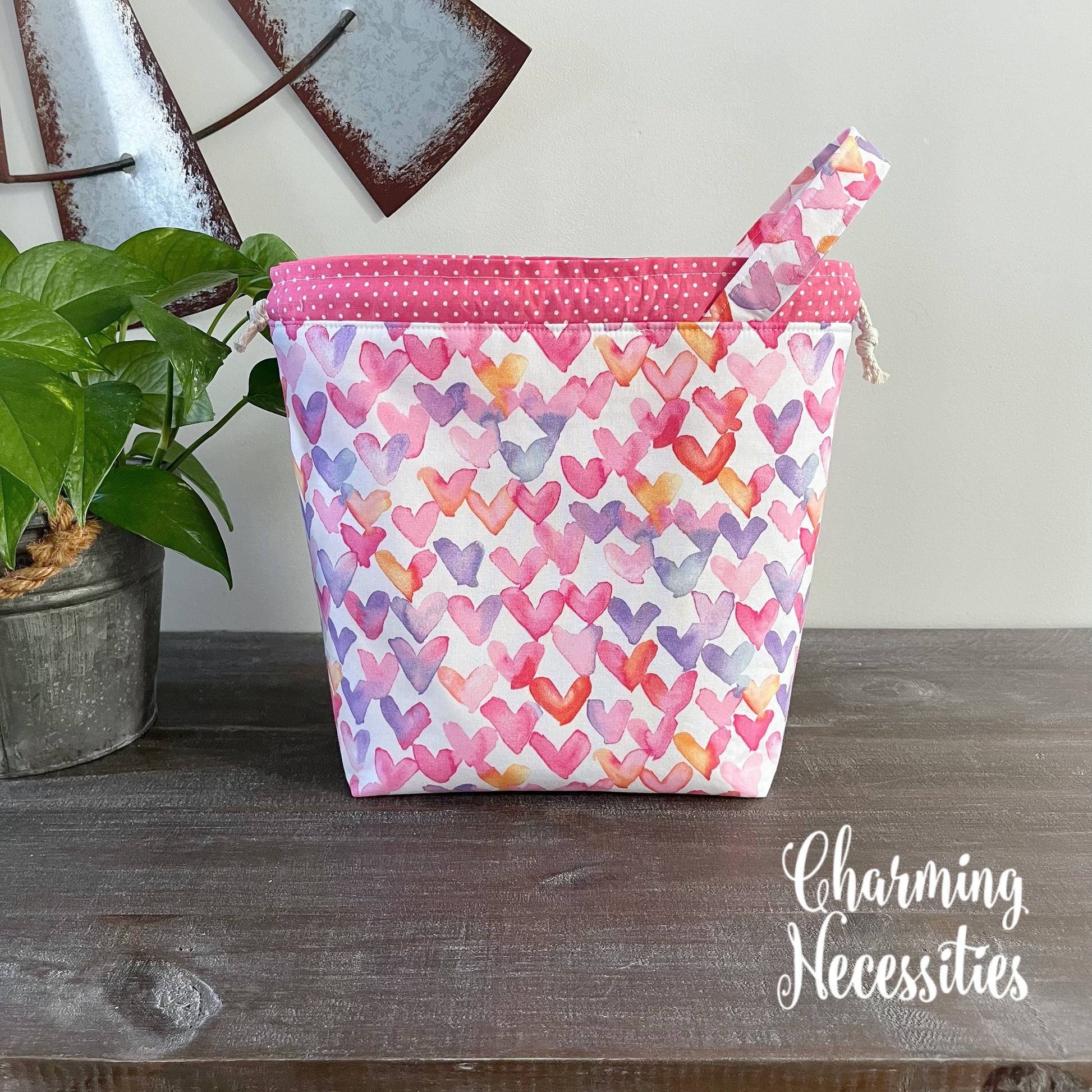 Colorful heart-patterned bag with pink top on a wooden surface, featuring 'Charming Necessities' branding.