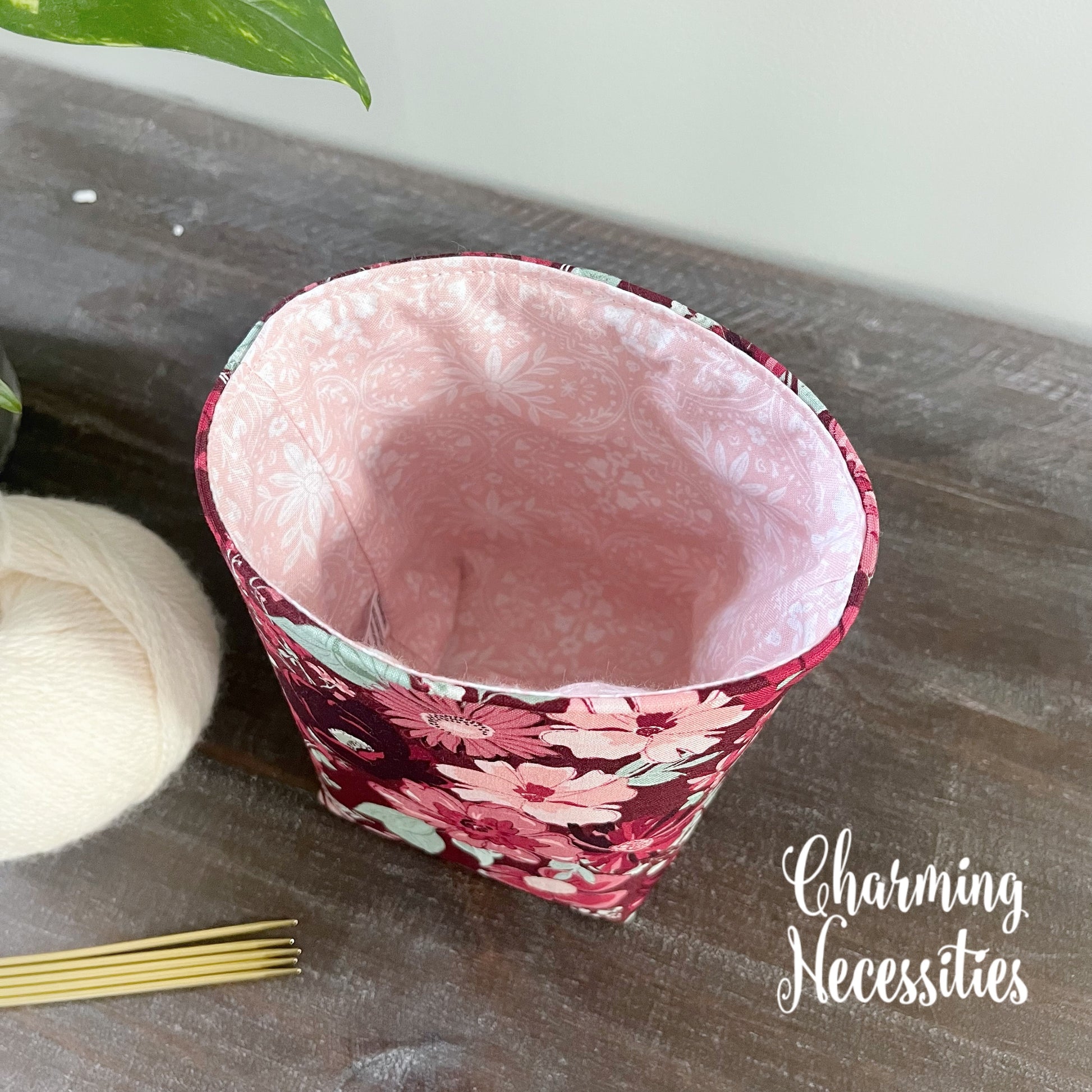 Yarn cozy with floral pattern on a wooden surface, next to yarn and knitting needles, with 'Charming Necessities' branding.