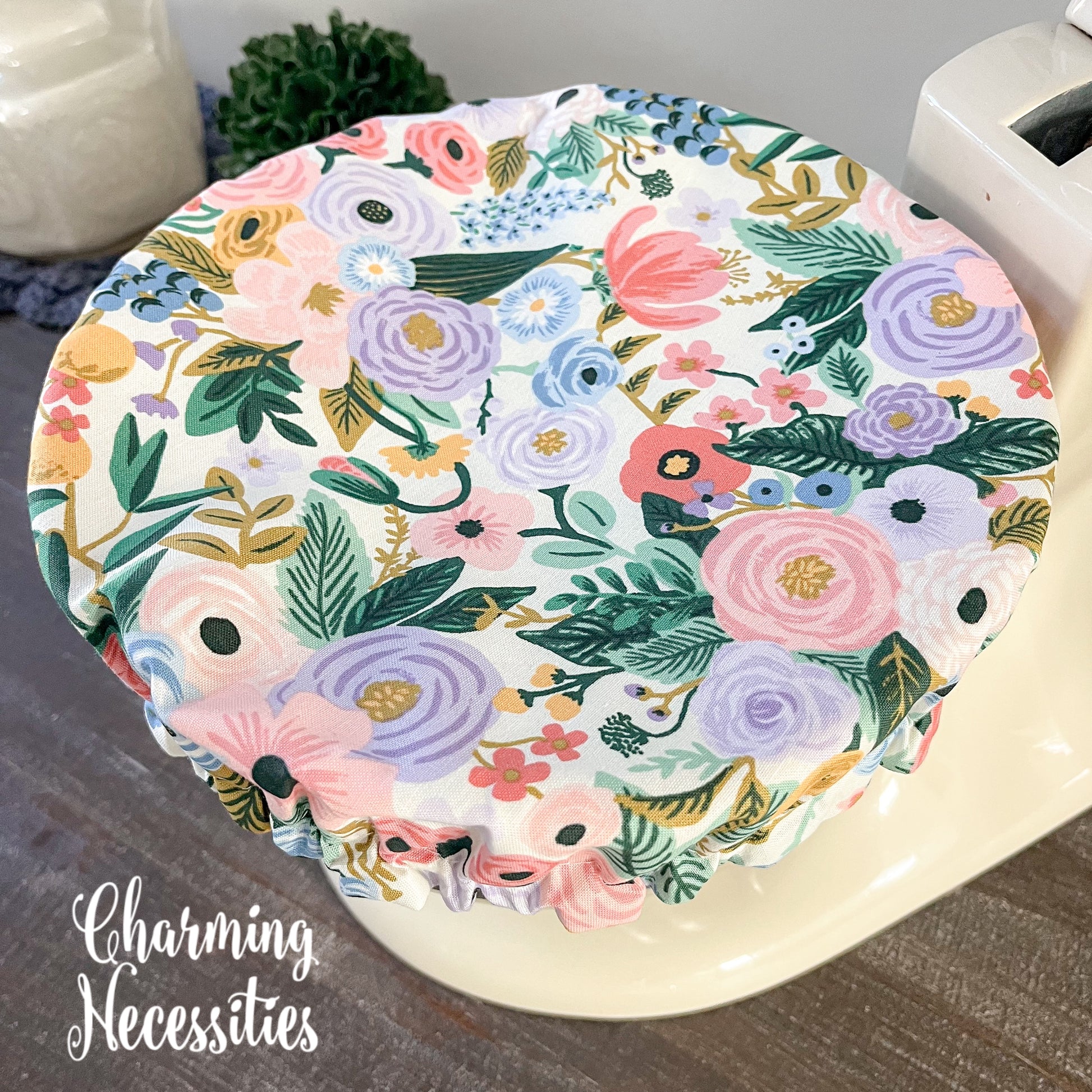 Floral-patterned bowl cover on a surface with 'Charming Necessities' branding.