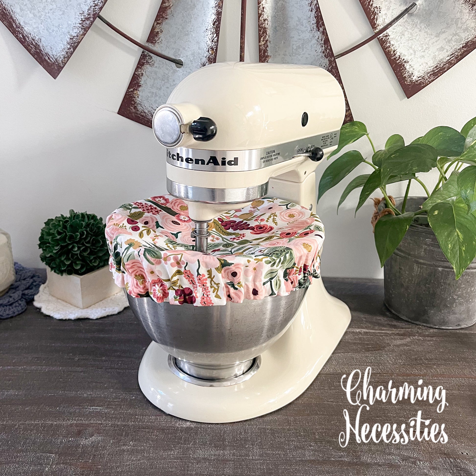 KitchenAid mixer with pink floral cover on a wooden surface, surrounded by decorative elements.