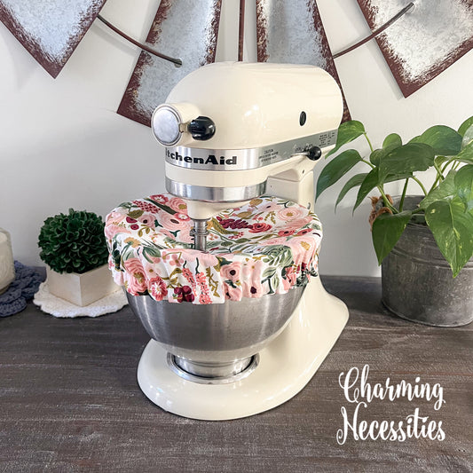 KitchenAid mixer with pink floral cover on a wooden surface, surrounded by decorative elements.