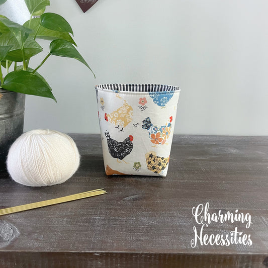 Yarn bowl with chicken pattern on a wooden surface next to yarn and knitting needles, with 'Charming Necessities' branding.