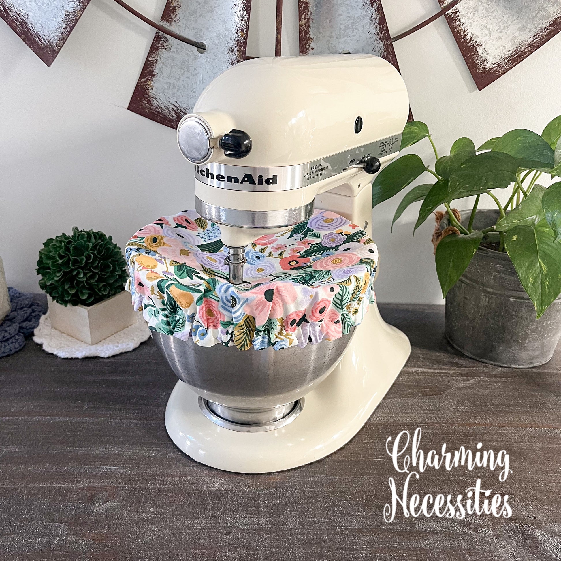 KitchenAid mixer with a floral cover on a wooden surface, surrounded by plants.