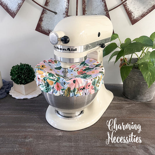 KitchenAid mixer with a floral cover on a wooden surface, surrounded by plants.