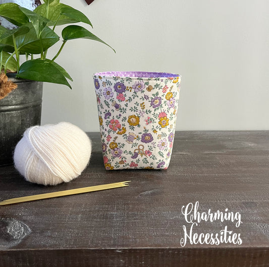 Fabric  yarn basket with floral pattern on a wooden surface next to yarn and knitting needles, with 'Charming Necessities' branding.